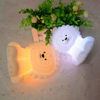 Two bear-shaped night lights, one yellow and one white, on a light gray surface with a plant in the background.